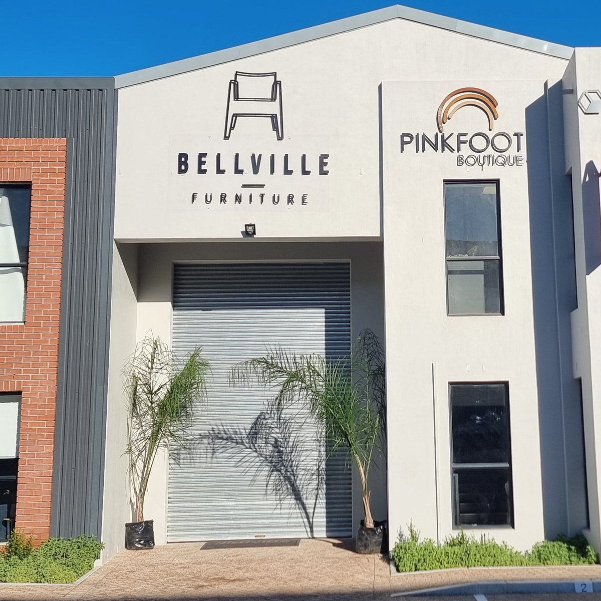 Front exterior of a modern industrial-style building with three main storefronts labeled Bellville Furniture, Pinkfoot Boards, and Deftco, under a clear blue sky with parked cars in the foreground.