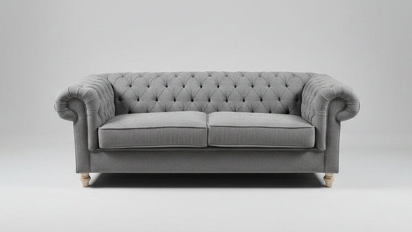 A gray tufted loveseat sofa with patterned throw pillows, styled in a showroom with a distressed blue-gray area rug, a small round coffee table, and a mustard yellow armchair.