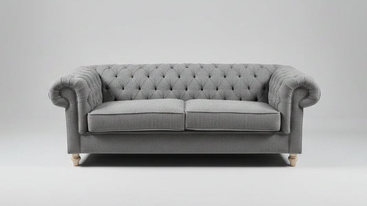 A gray tufted loveseat sofa with patterned throw pillows, styled in a showroom with a distressed blue-gray area rug, a small round coffee table, and a mustard yellow armchair.