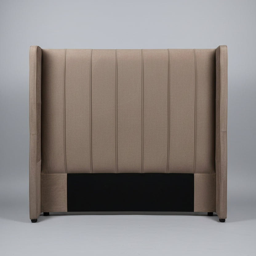 A tall upholstered headboard in taupe-brown fabric with vertical channel tufting and wingback sides, shown leaning against a light gray wall on a tiled floor.