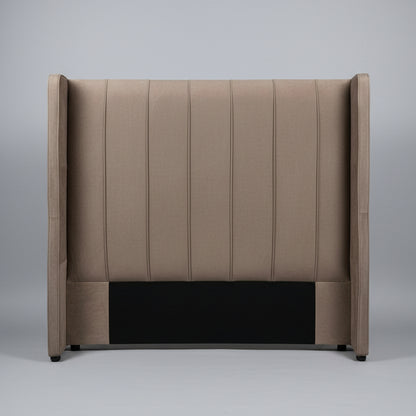 A tall upholstered headboard in taupe-brown fabric with vertical channel tufting and wingback sides, shown leaning against a light gray wall on a tiled floor.