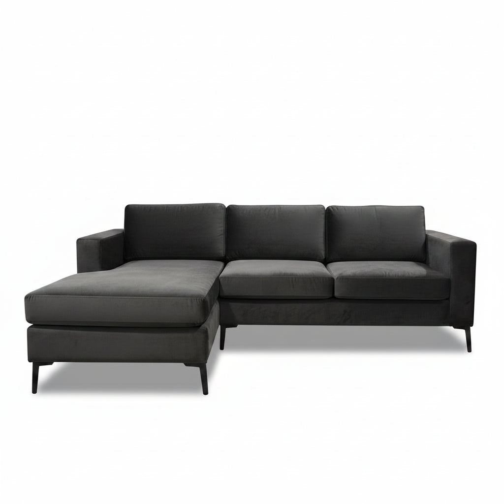 A dark gray velvet sectional sofa with an ottoman and a tropical print throw pillow, set in a furniture showroom with a round wooden coffee table in the foreground.