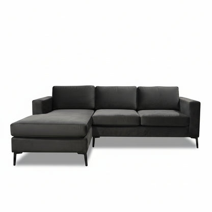 A dark gray velvet sectional sofa with an ottoman and a tropical print throw pillow, set in a furniture showroom with a round wooden coffee table in the foreground.