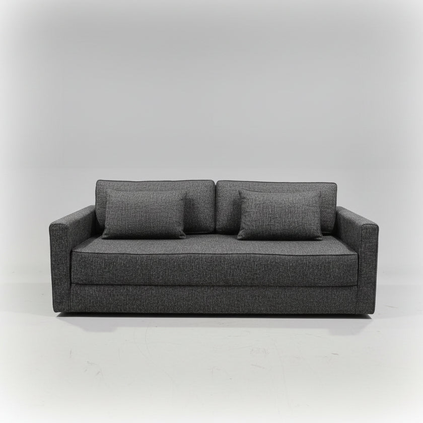 A dark gray upholstered loveseat sofa with two matching throw pillows, photographed in a furniture showroom with other home goods and displays visible in the background.