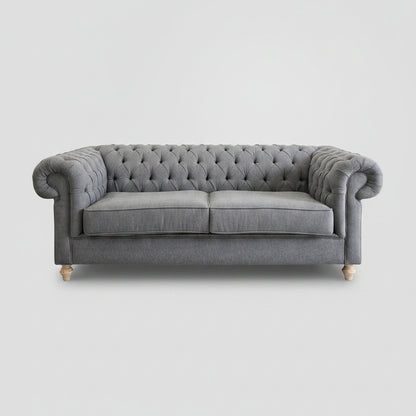 A gray tufted Chesterfield-style sofa with rolled arms and light wood legs, placed in a room with a white brick wall and a large abstract painting above it.