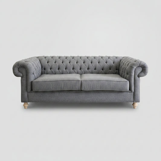 A gray tufted Chesterfield-style sofa with rolled arms and light wood legs, placed in a room with a white brick wall and a large abstract painting above it.