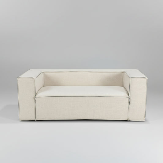 A light beige upholstered loveseat sofa in the foreground, photographed in a furniture showroom with other sofas and beds visible in the background.