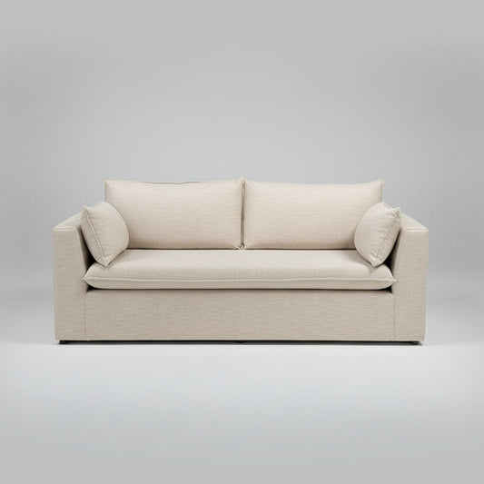 A light beige upholstered loveseat with two back cushions and two throw pillows, photographed in a furniture showroom with other sofas in the background.