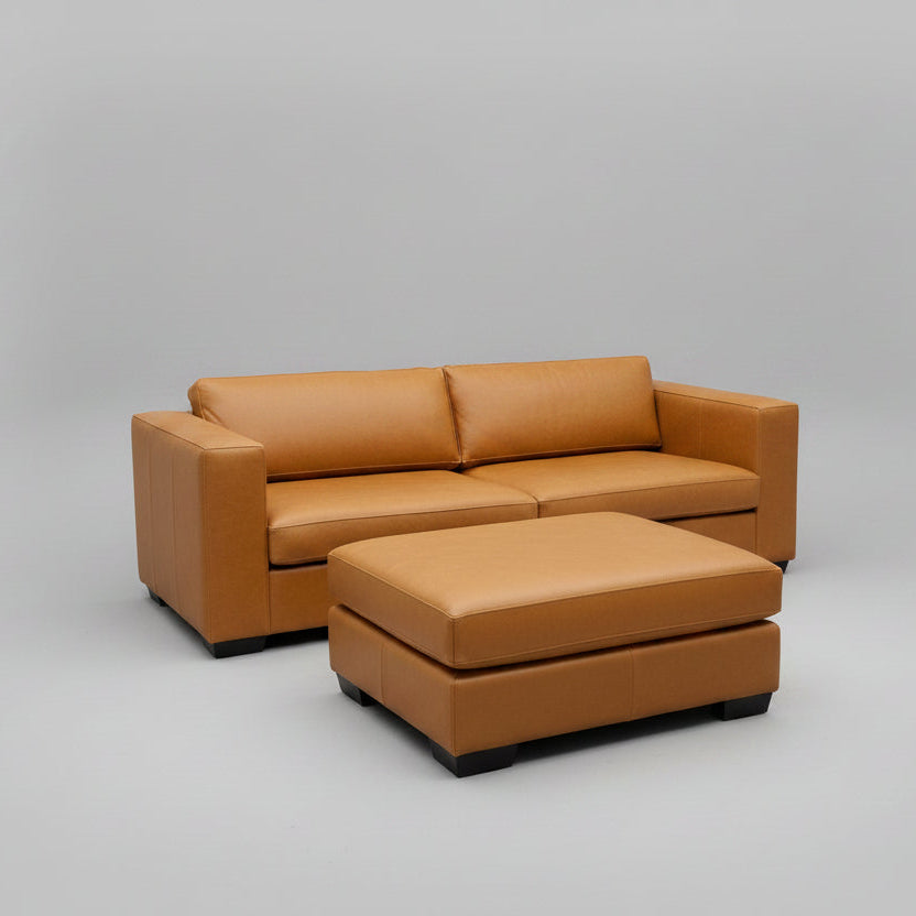 A modern living room setup featuring a tan leather two-seater sofa and a matching square ottoman on a gray patterned area rug. The room includes a dark gray accent wall, a small wooden side table with books and a plant, and a coat rack with garments.